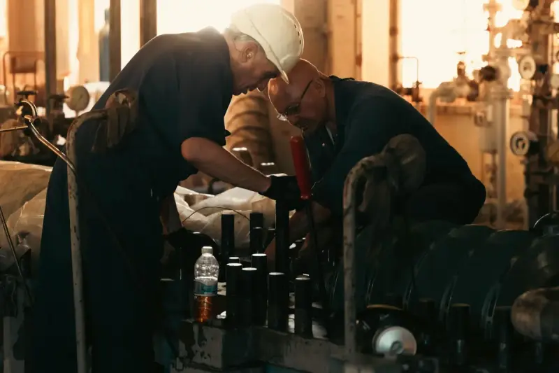 Two technicians in hard hats working together on plant machinery in a dim pump room