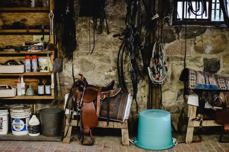 Western saddle on a stand beside bridles and tack hanging on a stone stable wall