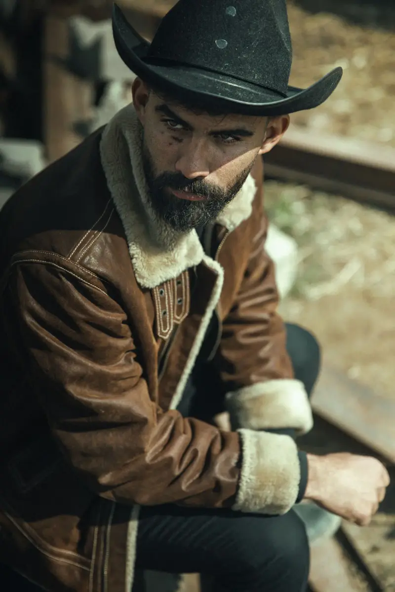 Bearded cowboy in a black hat and shearling-collar leather jacket seated by a corral