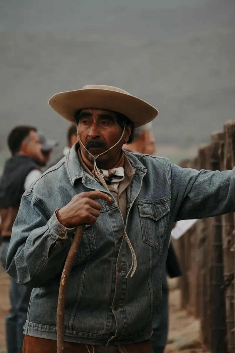 Older rancher in a wide-brim hat and worn denim jacket holding a rope at a corral