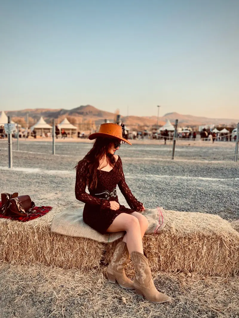 Woman in a rust cowboy hat, burgundy dress and tan western boots on a hay bale