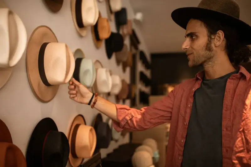 Man in a red shirt reaching for a hat on a wall of felt and straw hats in a hat shop