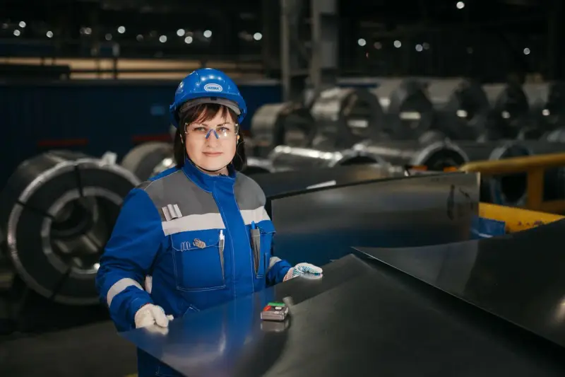 Woman in a hard hat and safety glasses inspecting sheet metal beside steel coils