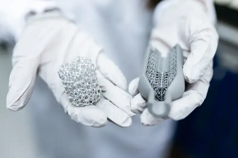 Gloved hands holding two 3D printed metal parts with fine lattice structures