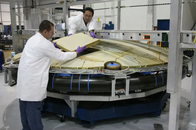 Two technicians in lab coats fitting a large panel onto a spacecraft component