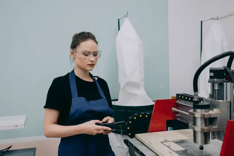 Woman in safety glasses and apron checking a handheld controller beside a CNC machine