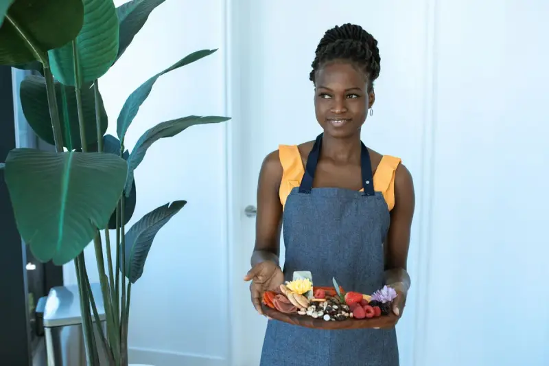Smiling woman in a denim apron holds a wooden charcuterie board with cured meats, cheese, berries and flowers