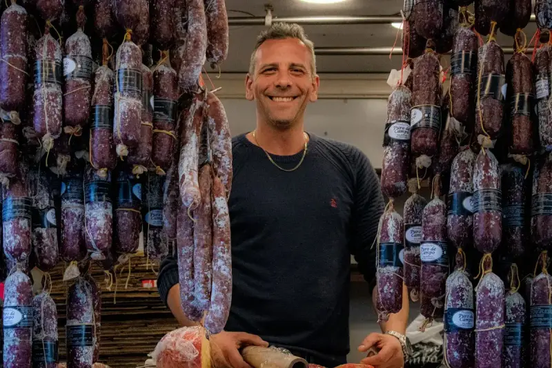 Smiling vendor stands between rows of hanging salami at a cured meat stall in an Argentine market