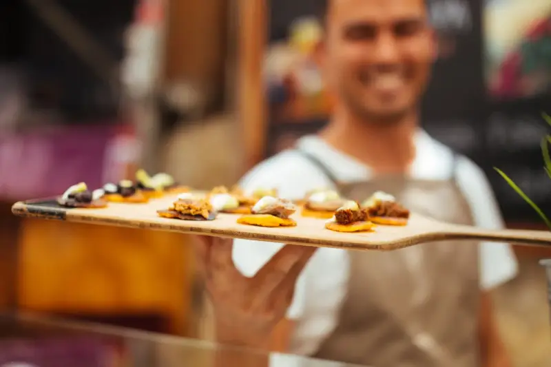 Smiling server in an apron presents a wooden paddle of gourmet canapés, with the bites in sharp focus