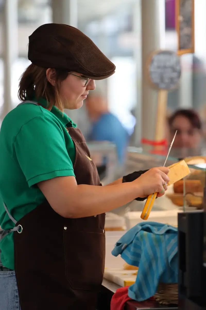 Woman in a cap and brown apron slicing a block of cheese at a market counter