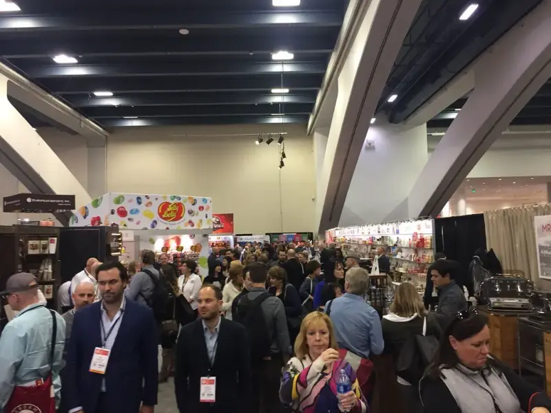 Buyers walking a Winter Fancy Food Show aisle lined with candy and snack exhibits
