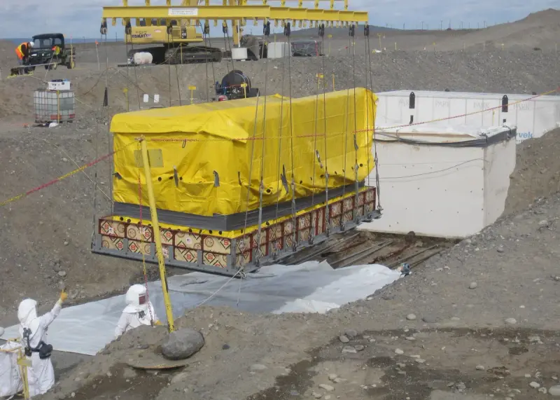 Workers in protective gear lifting a large wrapped box of transuranic waste