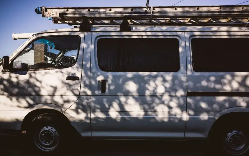 White work van with an extension ladder on its roof rack, dappled with tree shadows