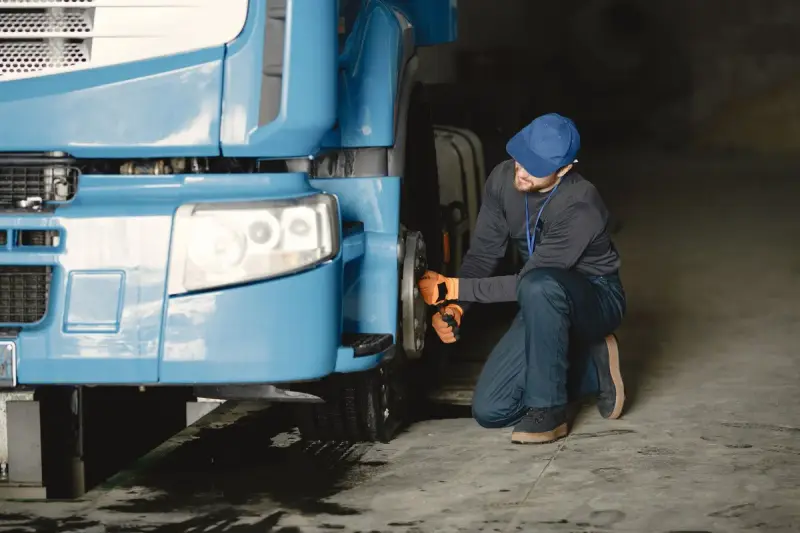 Mechanic in a blue cap and gloves kneeling to work on the front wheel of a blue truck