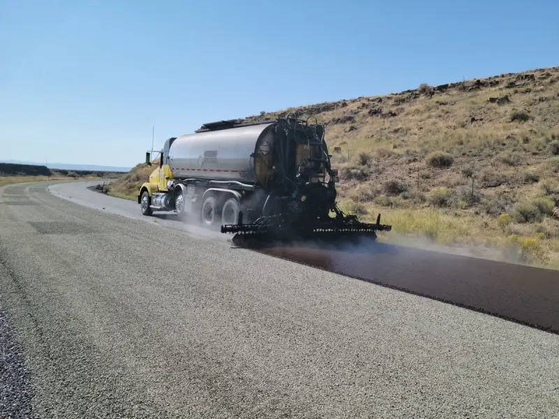 Distributor truck spraying a liquid asphalt coat across a rural road in dry hills