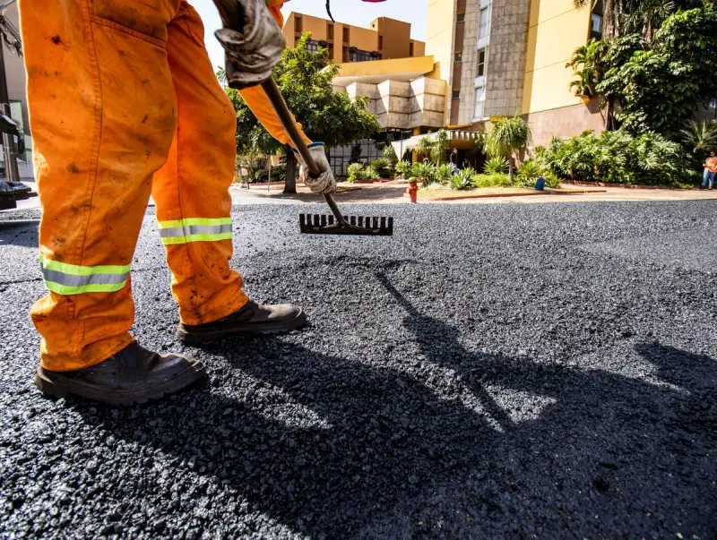 Worker in orange high-visibility gear raking hot asphalt mix on a city street