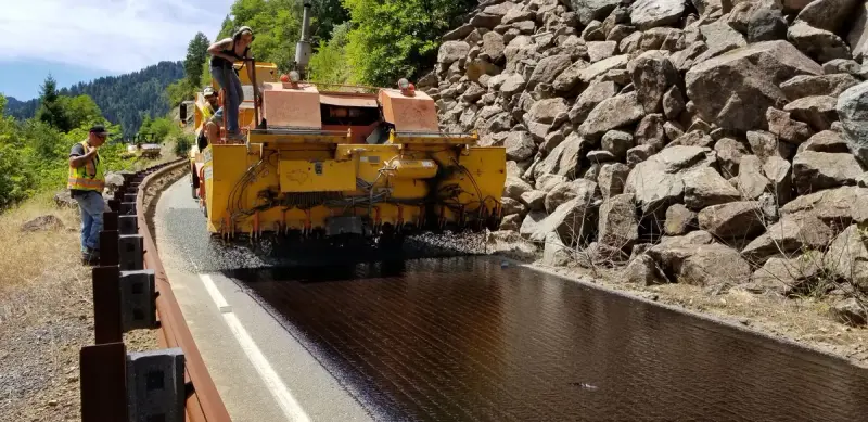Yellow paver screed laying fresh asphalt beside a guardrail on a rocky mountain road