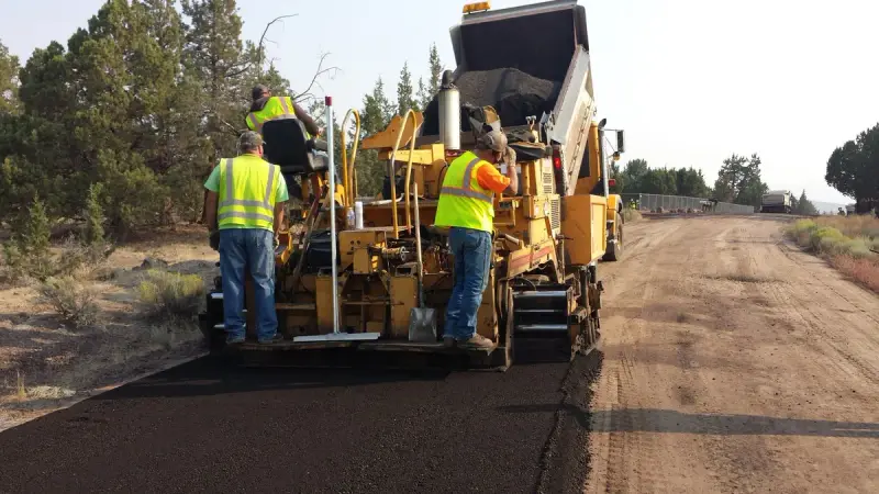 Two workers in high-visibility shirts riding and guiding an asphalt paver on a gravel road