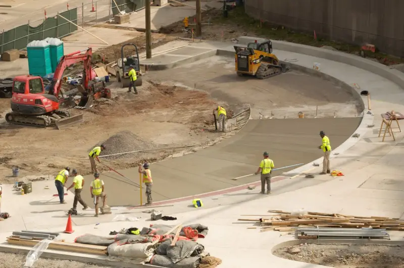 Crew in safety vests placing and finishing a long curved concrete pour with a compact excavator nearby