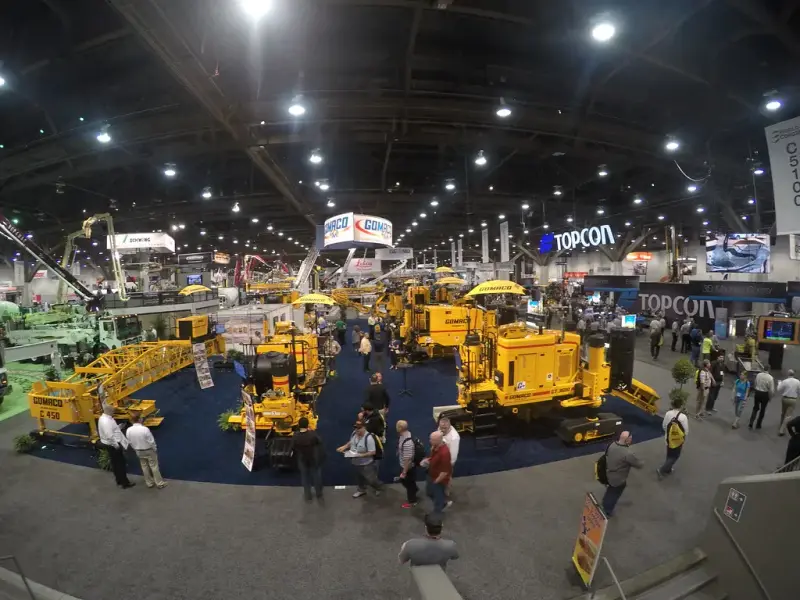 Overhead view of yellow slipform paving machines and visitors in a large convention hall