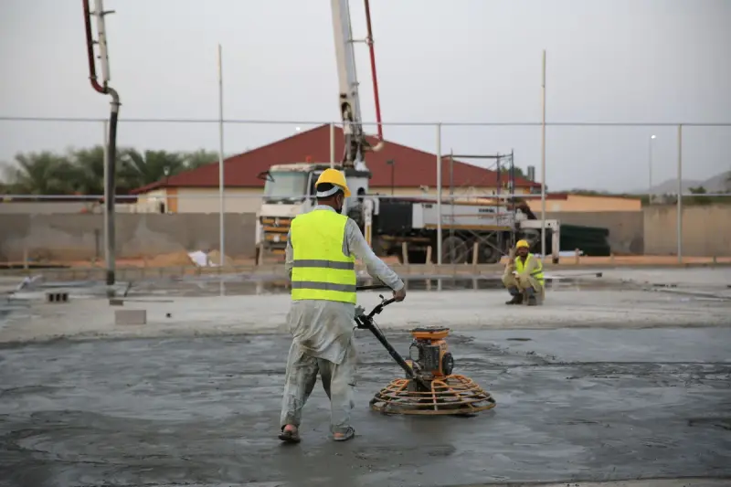 Worker in a hi-vis vest guiding a power trowel across a fresh concrete slab as a pump boom rises behind