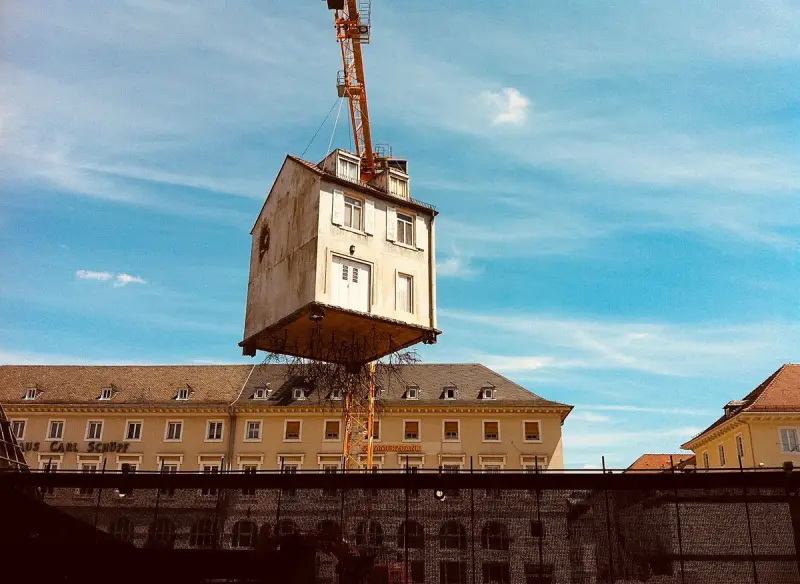 A small house-shaped building unit hanging from a crane against a blue sky