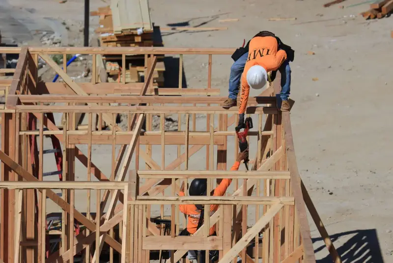 Two construction workers framing a wooden structure outdoors, one handing a tool to the other