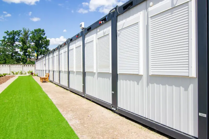 Row of metal-clad modular units with roller shutters beside a lawn and gravel path