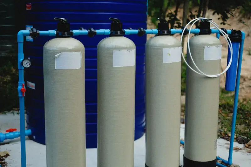 Four beige filter tanks connected by blue piping with a pressure gauge