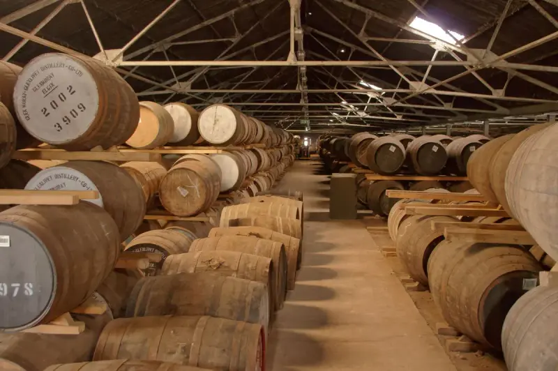 Long aisle between racks of stacked oak whisky casks inside a distillery aging warehouse