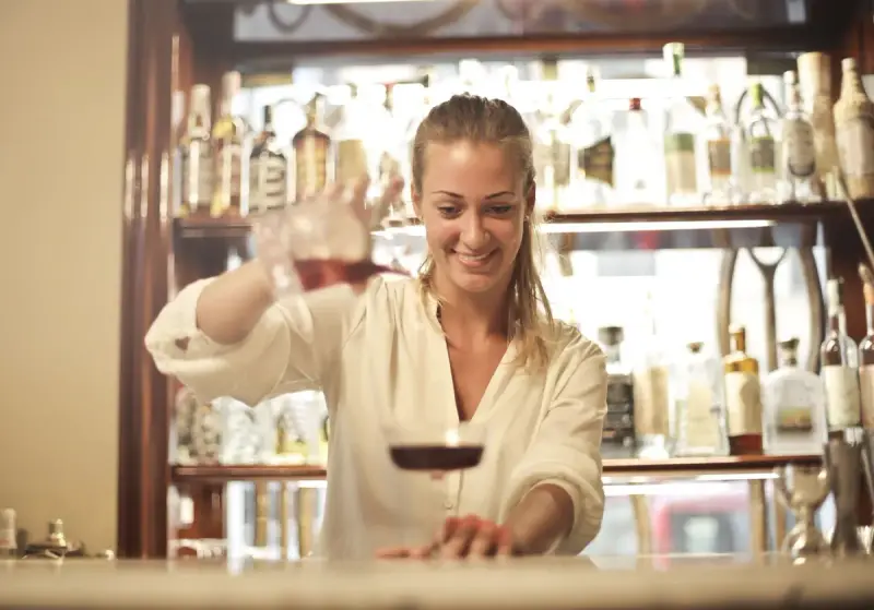 Smiling woman bartender pouring a red drink into a coupe glass at a bar counter