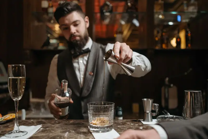 Bartender in a vest and bow tie measuring a spirit with a jigger into a glass
