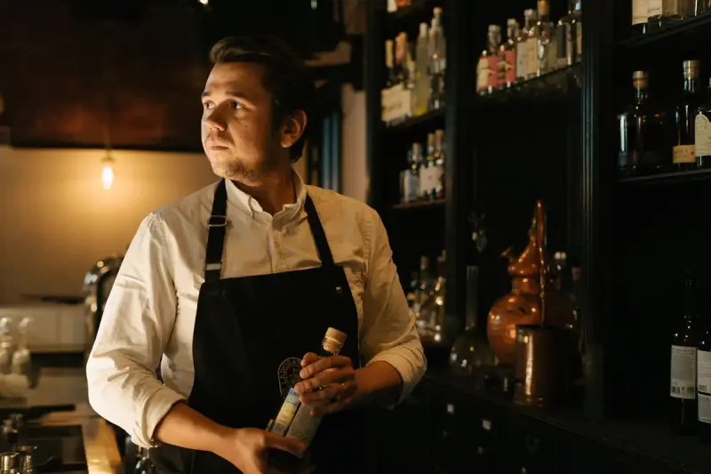 Bartender in a dark apron holding a bottle beside shelves of spirits and a copper still