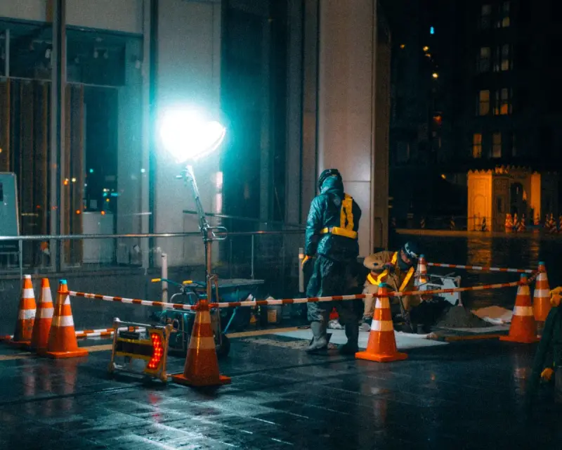 Worker in rain gear inside a coned-off street work zone lit by a tower light at night