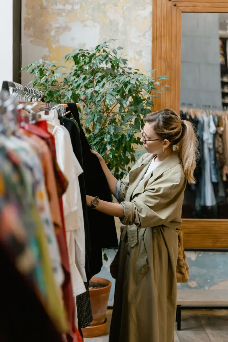 Woman in a khaki trench browsing a colorful clothing rail beside a mirror and plants