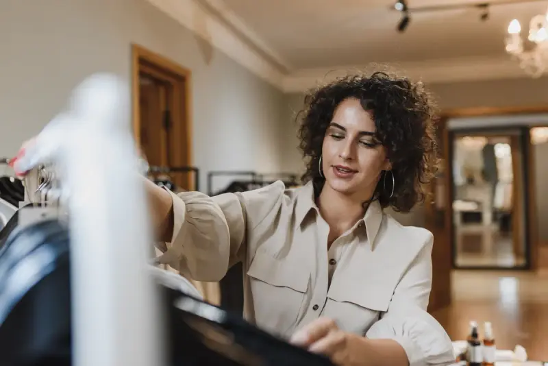Curly-haired woman in a cream shirt sorting garments on a rail in a bright boutique