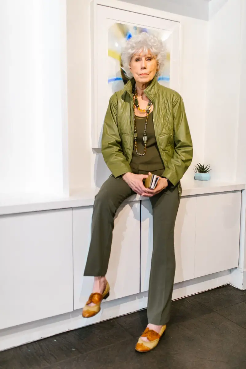Silver-haired woman in an olive jacket and trousers with tan loafers in a white gallery room