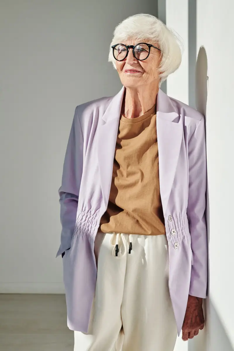 Older woman in round glasses, a lilac blazer, camel top and cream trousers