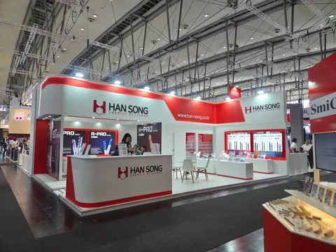 Han Song trade show booth built by Purple Exhibits: Wide view of a red, white and grey Han Song island booth with a tall header fascia, backlit product wall panels showing cutting tools and a white reception counter, in