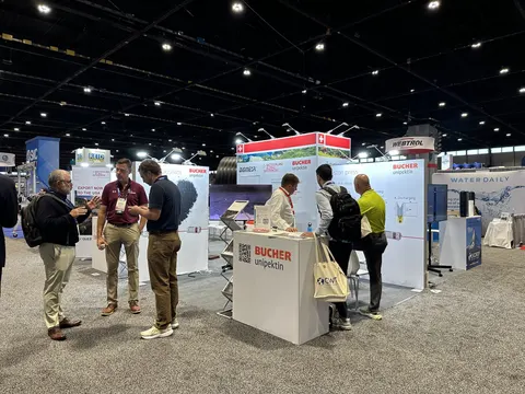Bucher Unipektin trade show booth built by Purple Exhibits: Busy inline booth for Bucher Unipektin with a red and white hanging sign, white counter with logo and several visitors talking in a large hall.
