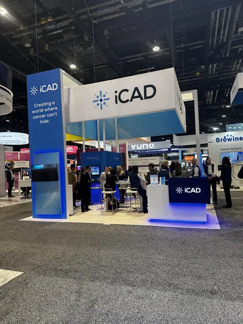 iCAD trade show booth built by Purple Exhibits: Blue and white iCAD island booth with a tall branded tower, overhead header sign, backlit demo screens, white counter with blue LED underglow and a crowd of visitors.