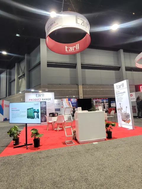 Taril trade show booth built by Purple Exhibits: Open Taril island booth on red carpet with a large round red-and-white hanging sign, freestanding lightbox panels about transformers, a white counter, chairs and potted pl