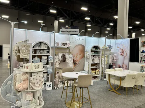 Piccolo Bambino trade show booth built by Purple Exhibits: Soft white and cream inline booth for Piccolo Bambino with arched product shelving, large baby-photo graphics, marble bistro tables and gold-legged velvet chairs