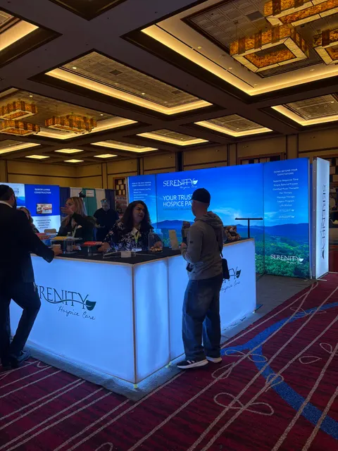 Serenity Hospice Care trade show booth built by Purple Exhibits: Blue mountain-landscape backlit wall and glowing white lightbox counters for Serenity Hospice Care inside a hotel ballroom, with staff behind the counter a