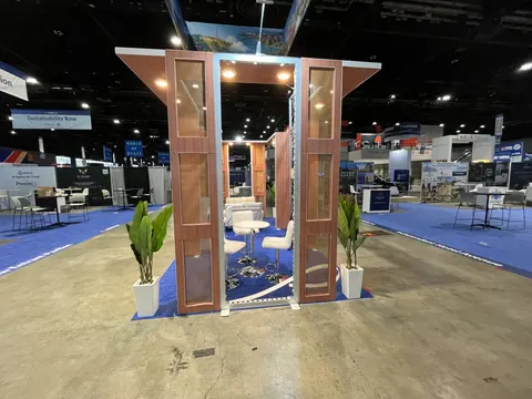 Trade show booth built by Purple Exhibits: Wood-slat portal entrance with copper-toned panels, potted palms, white bar stools and a blue carpet edge, photographed straight on with the wide show floor behind it.