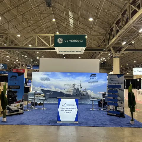 4 Marine Services LLC trade show booth built by Purple Exhibits: Inline backwall with a full-width naval warship photo, a small branded counter reading USA Caribbean Bridgeway to LATAM, bar stools, a brochure rack and fa
