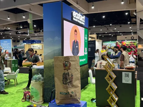 ecotact trade show booth built by Purple Exhibits: Ecotact coffee-packaging booth with a tall green field-scene graphic tower topped by an LED screen, kraft-paper bags on display, green turf flooring and staff serving at