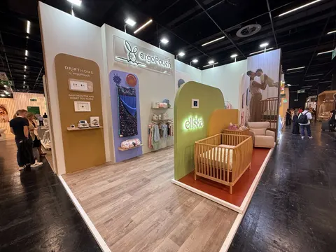 ergoPouch trade show booth built by Purple Exhibits: Warm cream ergoPouch baby-products booth with arched olive, terracotta and lilac niches displaying sleep bags, a wooden cot with a nursing chair on a rust rug, and woo