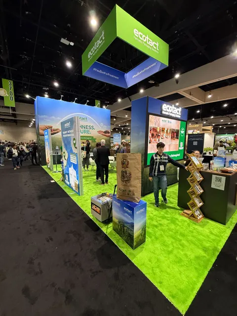 Ecotact trade show booth built by Purple Exhibits: Ecotact island booth with bright green turf flooring, a green-and-blue hanging sign, backlit nature graphics, a dark counter with a wooden literature rack and freestandi