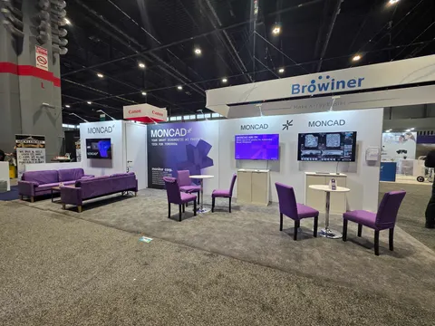 Moncad / Browiner trade show booth built by Purple Exhibits: Moncad inline booth with white walls, three wall-mounted screens showing medical imaging products, purple sofas and purple upholstered chairs at tall tables on